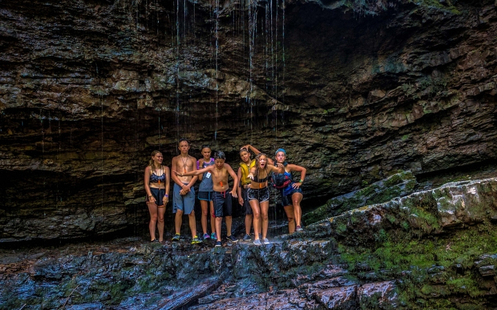 A group of young people wearing swimwear stand on a rocky ledge under a slowly trickling waterfall. 